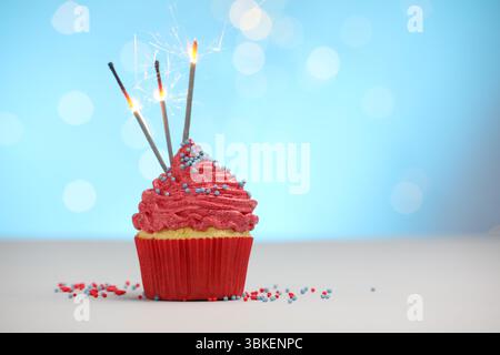 Burning sparklers in cupcake and sprinkles on white table against light blue background with blurred lights, space for text Stock Photo