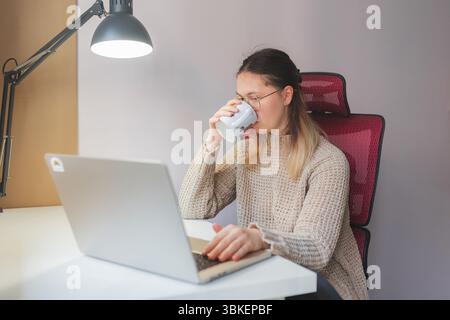 Woman drinking from mug while working from home on laptop. Red mesh chair, beige knit sweater, and focused gaze toward screen as she sips from white m Stock Photo