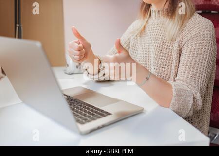 Woman gives double thumbs up during positive online meeting at home desk.Smiling woman shows two thumbs up in front of laptop, casual and warm outfit, Stock Photo