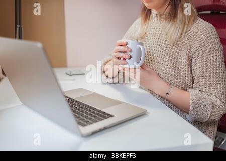 Woman holds mug while seated at desk with open laptop during work break. Close-up of woman’s hands around white coffee mug, resting casually near lapt Stock Photo