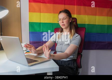 Serious woman raises hands during thoughtful online discussion near rainbow flag. Glasses-wearing woman in grey top communicates during remote meeting Stock Photo