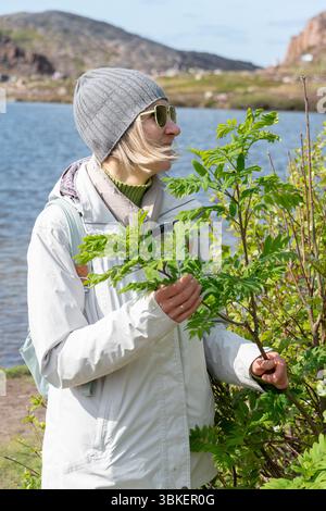 Woman in sunglasses and gray beanie stands by a green bush near a mountain lake, dressed in light outdoor clothing with a backpack Stock Photo