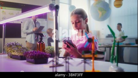 Talented Girl Sitting at Table and Using Smartphone, Conducting Biology Experiment on Plants with Tweezer and Magnifying Glass. Primary School Children During Lesson in Classroom. STEM Education. Stock Photo