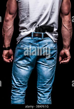 A man is seen from the back, wearing jeans and a white t-shirt. A handgun is tucked into the waistband of his jeans. The man's arms are muscular and h Stock Photo