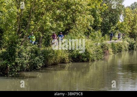 The FlowVélo long-distance cycle route (La Flow Vélo) runs for long stretches along the Charente River in the Nouvelle-Aquitaine region. Participants in the guided cycling tour by Die Landpartie ride along a bank path beside the water (possibly a canal parallel to the Charente), with tour guide Katell Nicoux in a high-visibility vest on the left. Trees and reeds line the bank at Rue de la Rivière, La Vallée, France. The route runs for around 400 kilometres from Sarlat-la-Canéda in the Périgord to the Île d'Aix. Stock Photo