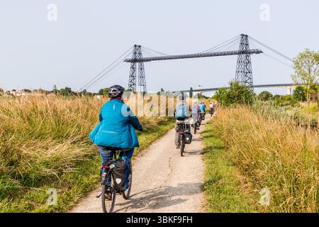 The transporter bridge (Pont transbordeur du Martrou) over the Charente River rises behind a cycle path on Rue Philibert Commerson, Rochefort, Nouvelle-Aquitaine, France. It is popular with cyclists and pedestrians. Participants in the guided cycling tour by Die Landpartie ride towards it in single file. Tall grasses line the path, and in the background on the right, the Martrou road viaduct (Viaduc de Martrou) of 1991 spans the river. The 68-metre-high steel pylons are visible from afar. Engineer Ferdinand Arnodin built the transporter bridge between 1898 and 1900. It links the town with Échi Stock Photo