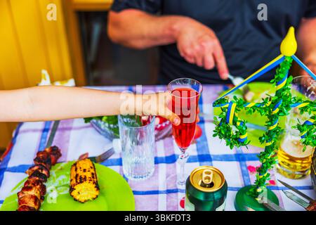 Child reaching for glass with red drink during Swedish Midsummer feast with grilled food and summer table setting. Sweden. Stock Photo
