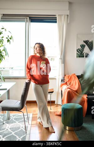Smiling young woman using mobile phone standing in living room at home. Stock Photo
