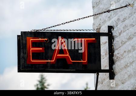 A glowing red neon sign displaying the word "EAT" mounted to a textured white brick wall with a blue sky in the background. Stock Photo