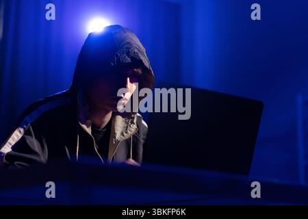 A young man in a black hoodie intently using a laptop in a neon-lit room, evoking themes of cybersecurity, hacking, coding, and digital privacy in a t Stock Photo