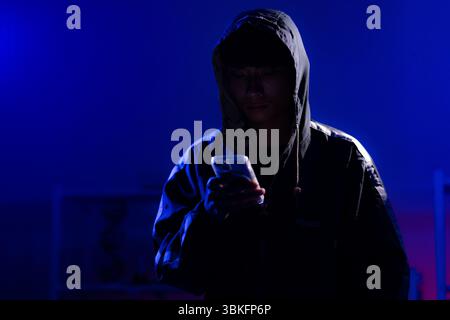 A young man in a black hoodie intently using a smart phone in a neon-lit room, evoking themes of cybersecurity, hacking, coding, and digital privacy i Stock Photo