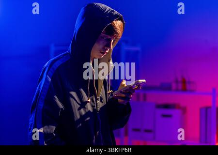 A young man in a black hoodie intently using a smart phone in a neon-lit room, evoking themes of cybersecurity, hacking, coding, and digital privacy i Stock Photo