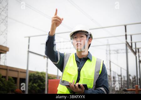A high voltage engineer is working attentively at substation wearing safety gear and pointing upwards while holding tablet in outdoor industrial envir Stock Photo