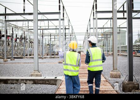 A two engineers are working at high voltage substation wearing safety helmets and reflective vests while inspecting electrical equipment in industrial Stock Photo