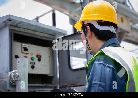 A power station engineer is working carefully inspecting electrical control panels wearing yellow safety helmet and reflective vest ensuring safety an Stock Photo