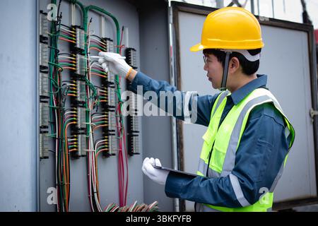 A power station engineer is working carefully on electrical wiring inside control panel wearing safety gear and yellow helmet showing focus and profes Stock Photo