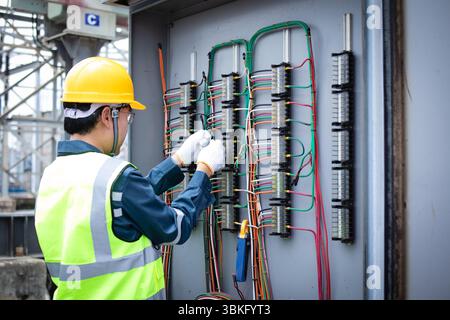 A power station engineer is working carefully on electrical wiring inside control panel wearing safety gloves and helmet showing focus and professiona Stock Photo