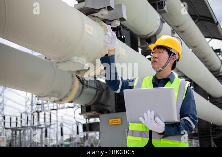 An engineer is working at power station inspecting large pipes while holding laptop and wearing safety gear showing focus and dedication in industrial Stock Photo
