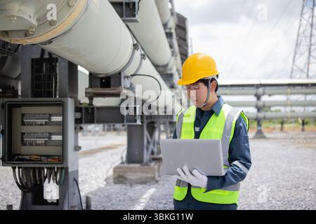 An engineer is working at power station inspecting large industrial pipes while using laptop to ensure efficient operation and safety in outdoor facil Stock Photo