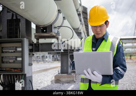 An engineer working at power station uses laptop for inspection and maintenance wearing safety helmet and reflective vest in industrial outdoor settin Stock Photo