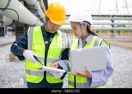 A two engineers are working together at power station outdoors with safety helmets and reflective vests while reviewing documents and using laptop in Stock Photo