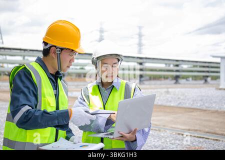 An engineer working at power station discussing project details on laptop outdoors with safety helmet and vest Stock Photo