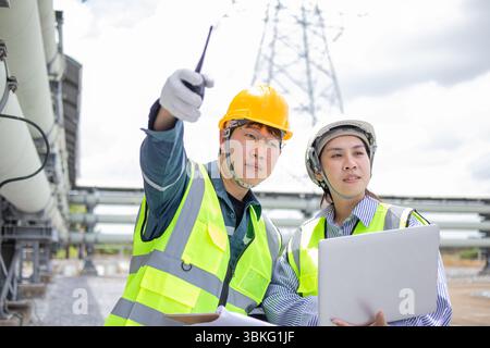 A two engineers working at power station are inspecting equipment with focus and determination while wearing safety helmets and reflective vests in in Stock Photo