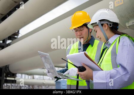 An engineer working at power station wearing safety helmet and reflective vest while checking laptop and clipboard with walkie talkie in industrial en Stock Photo