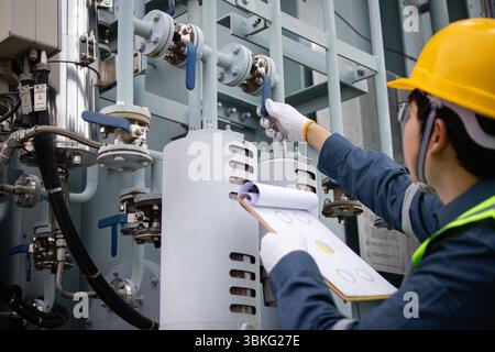 An engineer performs inspection on industrial equipment in substation checking valves and pipes while holding clipboard to ensure transformer system o Stock Photo