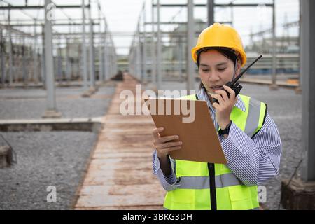 A power engineer wearing yellow helmet and reflective vest works at substation holding clipboard and communicating with staff using walkie talkie to e Stock Photo