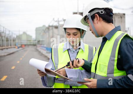 A two engineers work together at power substation with staff wearing safety helmets and reflective vests discussing plans and using walkie talkie for Stock Photo