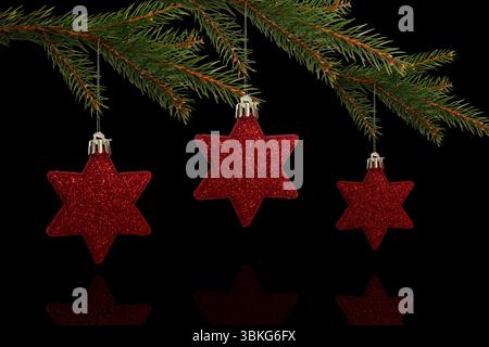 Three red glittery star ornaments are hanging on pine branch above black glossy surface, copy space Stock Photo
