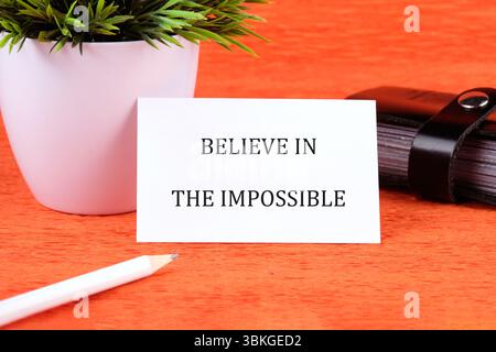 Motivational quote. BELIEVE IN THE IMPOSSIBLE text written on a card next to a small potted plant and stationery. Stock Photo
