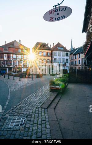 The setting sun shines between half-timbered houses on the Place de la Sinne with the Friedrich fountain in the old town centre of Ribeauvillé, Alsace Stock Photo