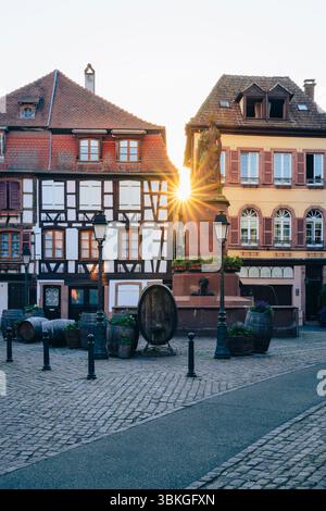 The setting sun shines between half-timbered houses on the Place de la Sinne with the Friedrich fountain in the old town centre of Ribeauvillé, Alsace Stock Photo