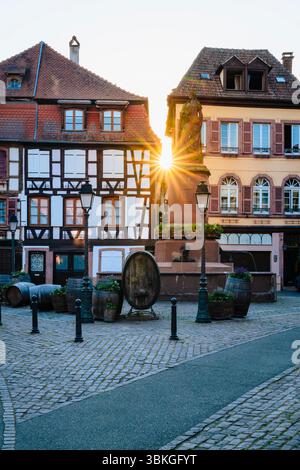 The setting sun shines between half-timbered houses on the Place de la Sinne with the Friedrich fountain in the old town centre of Ribeauvillé, Alsace Stock Photo
