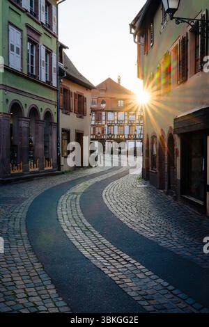 The setting sun shines between half-timbered houses through a winding alley in the old town centre of Ribeauvillé, Alsace Stock Photo
