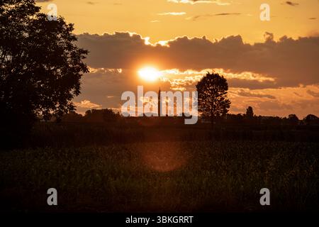 Sun setting behind a picturesque bell tower in the countryside, casting a warm glow over a corn field and silhouetted trees Stock Photo