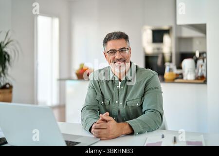 Happy middle aged man wearing glasses sitting with laptop at home table. Stock Photo