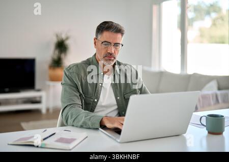 Serious older mature man wearing glasses using laptop computer at home table. Stock Photo