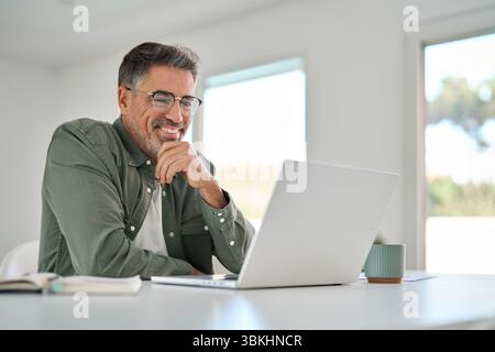 Happy older mature man using laptop computer at home table. Stock Photo