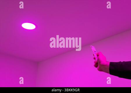 Hand holding a remote control to change the color of the light in the living room, purple led bulb on the ceiling Stock Photo