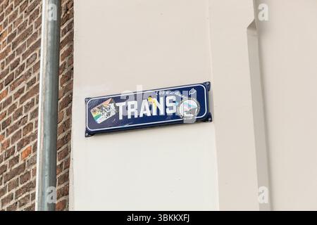A street sign that reads "TRANS" is mounted on a white wall next to a brick building. Stickers adorn the sign Stock Photo