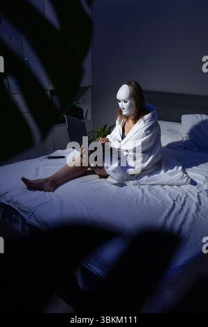 In a dimly lit room, a person draped in a blanket sits on a bed, using a laptop. Their face is obscured by a white mask, highlighting concerns over data security and fraud. Stock Photo