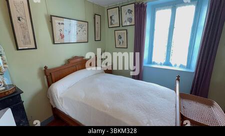 Bedroom with wooden bed, white linens, and framed artwork on light green walls. Window with blue frame and purple curtains. Stock Photo