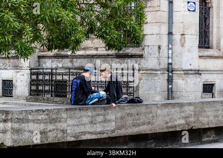 Everyday life in Angoulême: two young people sit on the wall outside the town hall – recognisable by the speech bubble lettering – and share a moment of digital togetherness. Comics and the present merge. Angoulême is recognized as a UNESCO Creative City in the category of literature since 2019. Rue de Genève, Angoulême, Nouvelle-Aquitaine, France Stock Photo