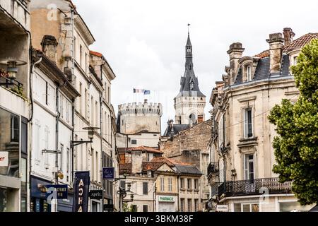 The town hall (Hôtel de Ville), with its tall bell tower and a crenellated medieval tower, rises above a picturesque street with historic buildings, Place Saint-Martial, Angoulême, Nouvelle-Aquitaine, France. The French and European flags fly from the crenellated tower. The bell tower carries a tall spire with a weather vane. Wrought-iron balconies decorate the façades, and shops line the ground floors, among them a pharmacy. Architect Paul Abadie built the Gothic Revival town hall from 1858 on the site of the former castle of the Counts of Angoulême, and it opened in 1870. Two towers of the c Stock Photo