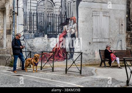 Man walking his dog while a woman sits on a bench using her mobile phone. In the background is a mural showing a couple embracing. Angoulême is recognized as a UNESCO Creative City in the category of literature since 2019. Rue Saint-André, Angoulême, Nouvelle-Aquitaine, France Stock Photo