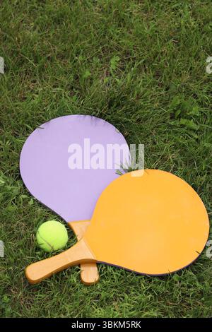 Beach tennis rackets and ball on grass, top view. Active lifestyle, games and sports. Beach tennis set. Bright wooden rackets for outdoor games. Summe Stock Photo