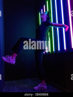 A woman in a dynamic pose standing in a dark room lit by vibrant neon lights, creating a striking contrast of colors and shadows. The image captures a Stock Photo